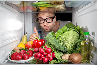 Ein Mann mit auffälliger Brille schaut in den Kühlschrank mit Gemüse und Obst.
