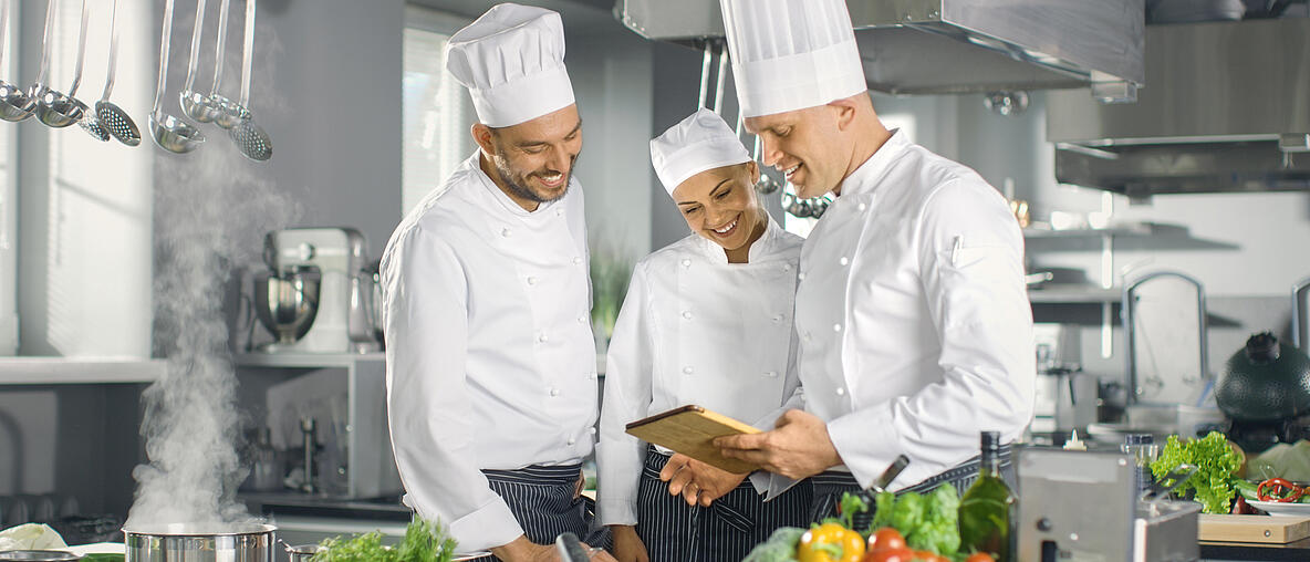3 Köche stehen in einer Küche und planen Essen an einem Tablet