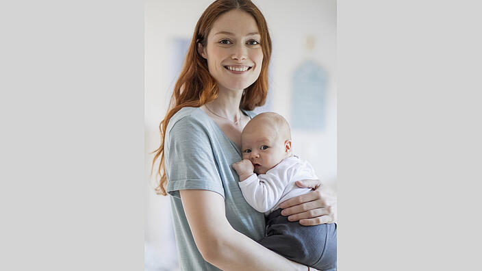Junge Frau hält ein Baby stehend auf dem Arm.