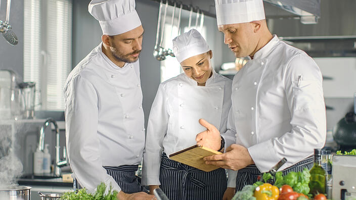 Küchensituation: zwei männliche Köche und eine webliche Köchin stehen zusammen mit Kochmützen und Schürze bekleidet und diskutieren miteinenader.