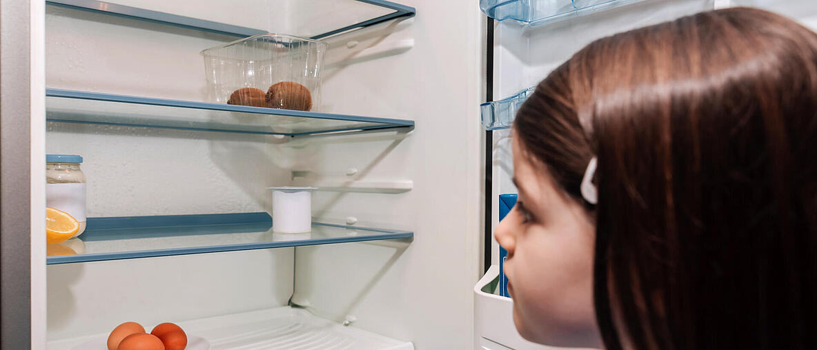  Ein Mädchen schaut in einen nahezu leeren Kühlschrank.  
