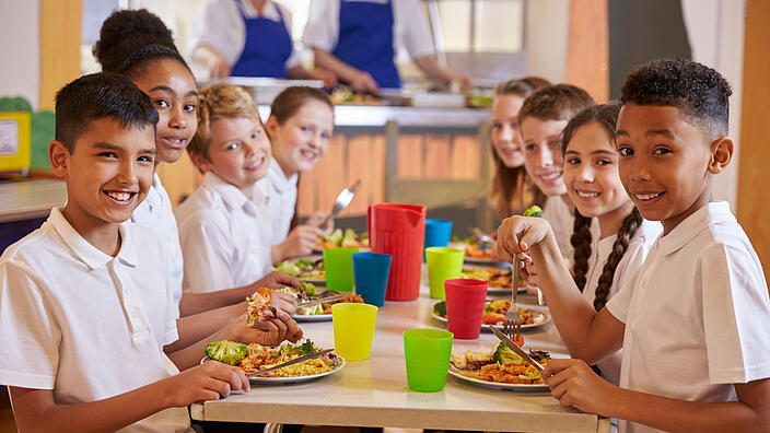 Kinder an einem Tisch in einer Grundschul-Cafeteria schauen in die Kamera