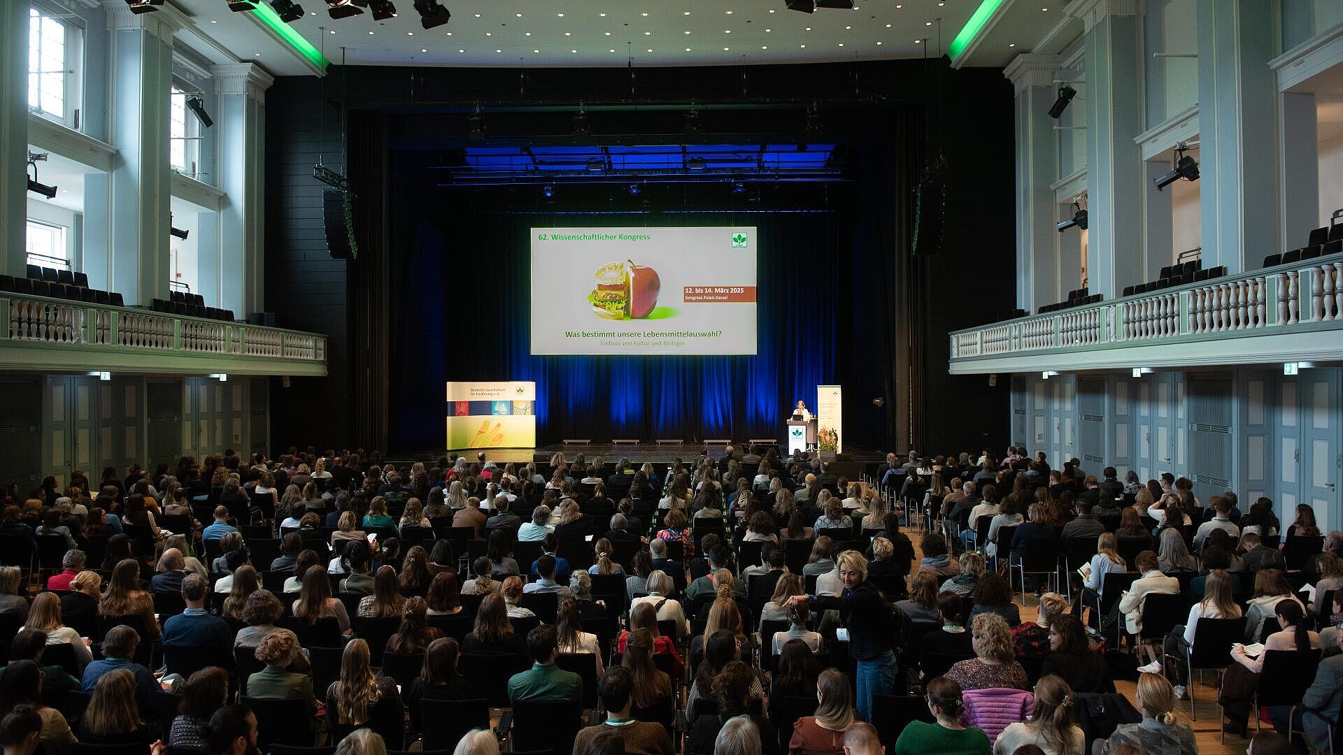 Vollbesetzter Festsaal mit Publikum, im Hintergrund die Bühne mit Präsentation