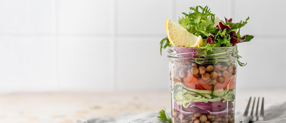 Salat mit Linsen und Gemüse in einem Glas mit Gabel auf Küchentisch.