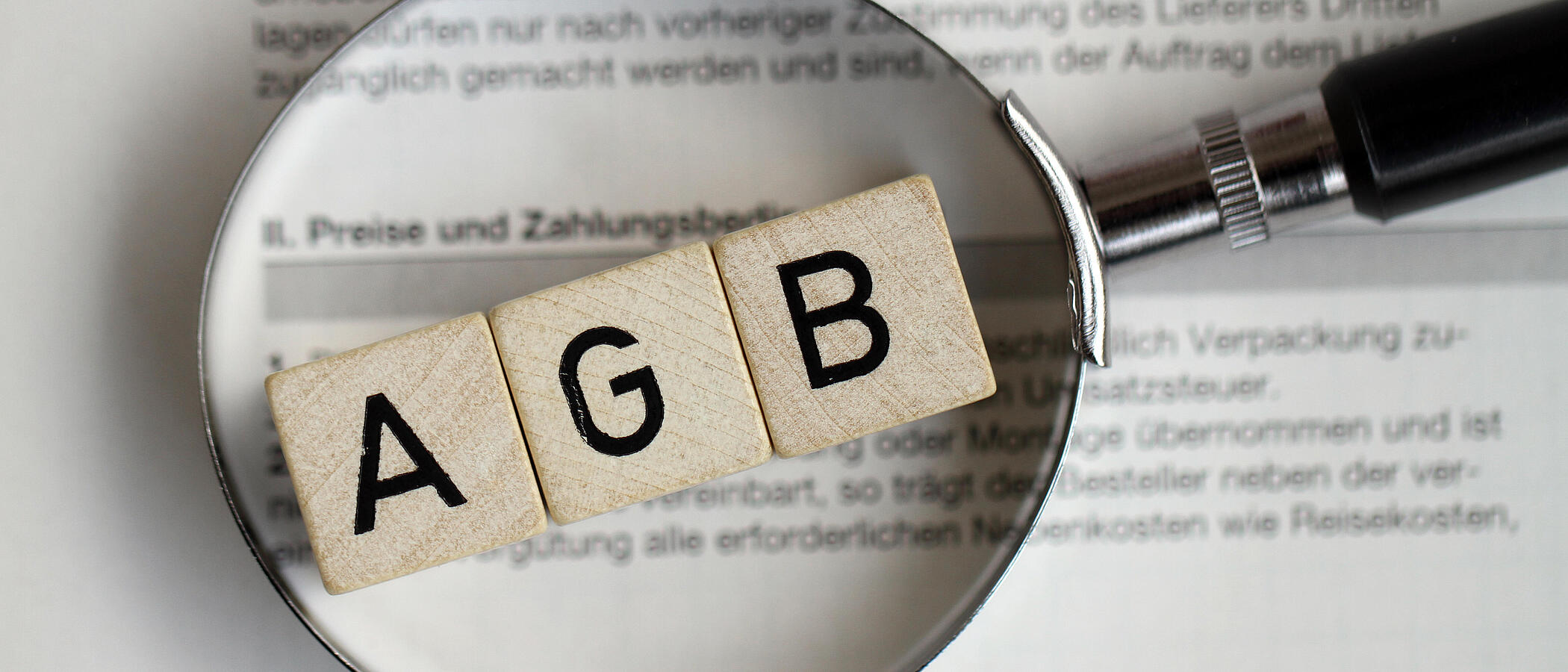 Unter einer Lupe erscheinen die Buchstaben AGB auf Holzwürfeln gedruckt. Sie liegen auf einem beschrifteten Dokument.
