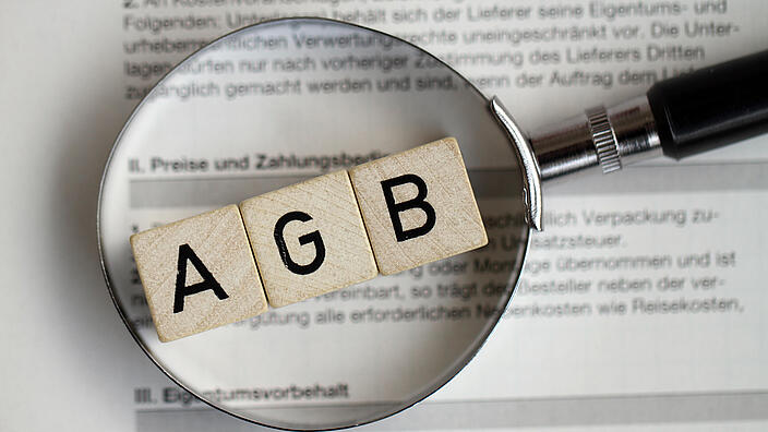Unter einer Lupe erscheinen die Buchstaben AGB auf Holzwürfeln gedruckt. Sie liegen auf einem beschrifteten Dokument.
